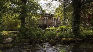 Una baita rustica con una veranda in legno e scale che portano a un portico, circondata da vegetazione lussureggiante e un ruscello.