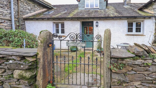 A stone cottage with a metal gate, a sign reading "Jane's Cottage," and a stone wall.