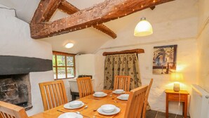 Comedor con una mesa de madera dispuesta para seis personas, rodeada de sillas de madera. Se observa una chimenea a la izquierda y una ventana con cortinas a la derecha.