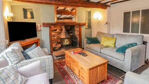 Una sala de estar acogedora con chimenea de piedra, vigas de madera, un sofá gris con cojines de colores, un sofá a cuadros y una mesa de centro de madera, además de un televisor de pantalla plana.