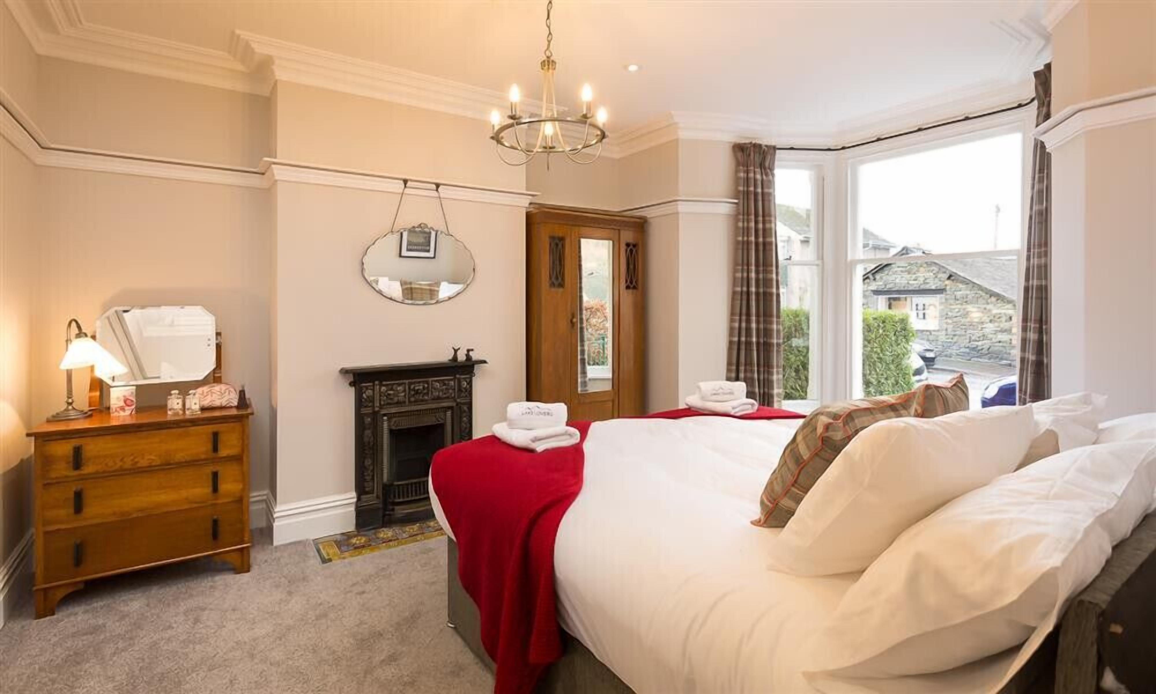 A hotel room with a bed, a wooden dresser, a mirror, a fireplace, and a window with a view of buildings.