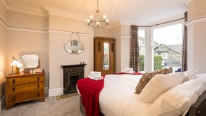 A hotel room with a bed, a wooden dresser, a mirror, a fireplace, and a window with a view of buildings. - Thirty Blencathra Street (Keswick)
