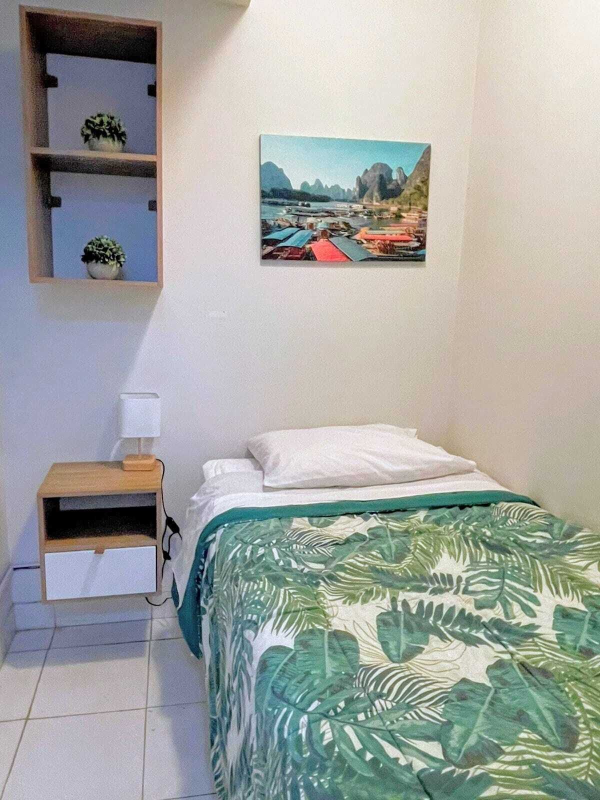 A bedroom with a bed, a nightstand, a shelf with plants, and a painting of a landscape.