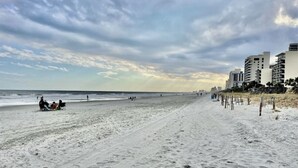 A wide sandy beach with footprints and a few people. There are buildings in the background.
