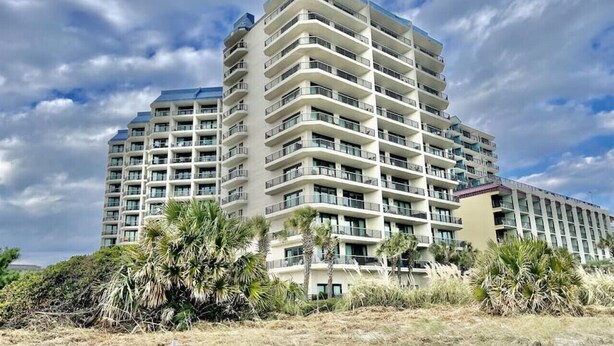 A multi-story hotel with balconies, located near a beach with sparse vegetation.