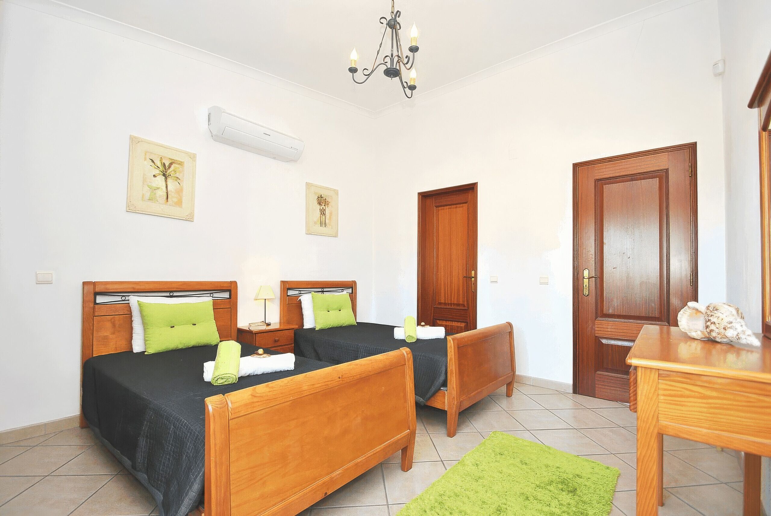 A hotel room with two wooden beds, a green rug, a wooden table, and a door.