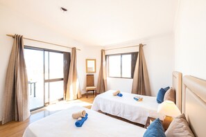A hotel room with two beds, a chair, a table, and large windows with curtains.