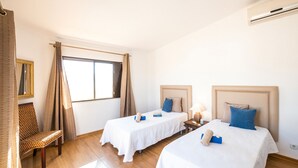 A hotel room with two beds, a chair, a window with curtains, and an air conditioning unit.