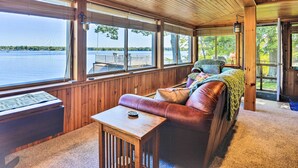 A cabin with a wooden interior, a leather couch, a table, and large windows overlooking a lake. - Waterfront Wellesley Island Getaway w/ Deck! (Wellesley Island)