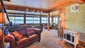 A spacious living room with a leather sofa, a fireplace, large windows, and a view of a body of water. - Waterfront Wellesley Island Getaway w/ Deck! (Wellesley Island)