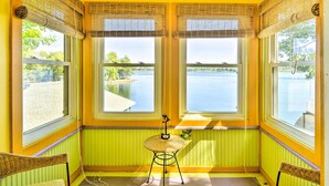 A room with two large windows overlooking a body of water, a wicker chair, and a small table with a decorative item. - Waterfront Wellesley Island Getaway w/ Deck! (Wellesley Island)