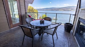 Varanda com mesa redonda e quatro cadeiras de vime, com vista para um lago com barcos e colinas ao fundo.