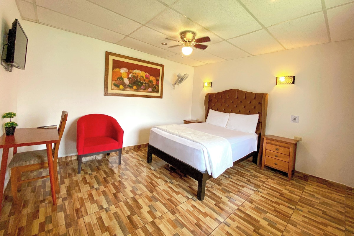 A hotel room with a bed, a desk, a chair, a ceiling fan, and a wall-mounted light fixture.