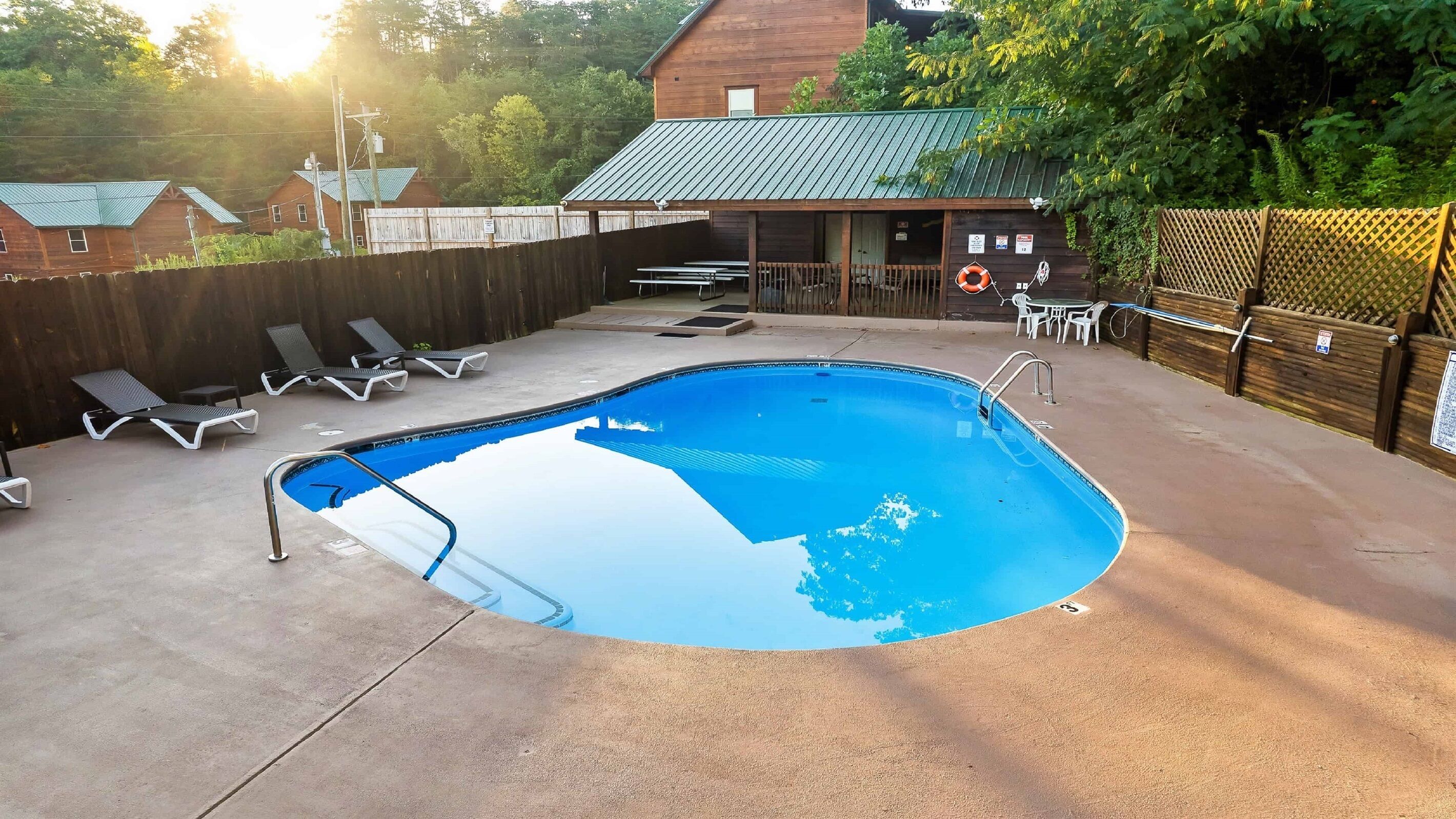 A swimming pool with lounge chairs, a covered seating area, and a wooden building in the background.