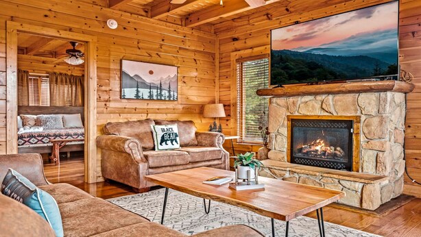 A cozy log cabin interior with a stone fireplace, a large TV mounted on the wall, and a comfortable seating area with a couch and a sofa.