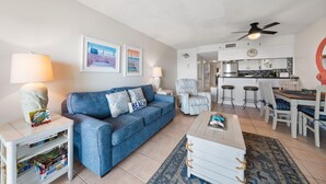 A living room with a blue sofa, a white coffee table, and a dining area with a wooden table and chairs. - Gulf Shores Condo Clearwater 8C | Sleeps 6 (Gulf Shores)