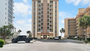 A multi-story hotel with a central vertical sign, a parking lot with a few vehicles, and palm trees around. - Gulf Shores Condo Clearwater 8C | Sleeps 6 (Gulf Shores)