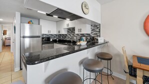 A modern kitchen with white cabinets, a stainless steel refrigerator, a black countertop, and a clock on the wall. - Gulf Shores Condo Clearwater 8C | Sleeps 6 (Gulf Shores)