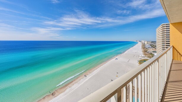 Blick auf einen Strand vom Balkon aus, mit blauem Himmel, sandigem Strand und einem Hotel im Hintergrund.