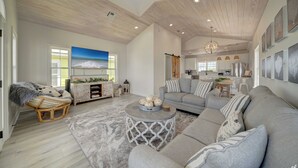 A spacious living room with a large gray sofa, a round coffee table, and a wicker chair. A kitchen is visible in the background with white cabinets and a hanging light fixture. - Summer Towne cottage w/ pool and beach access (Panama City Beach)