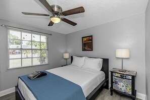A bedroom with a bed, a ceiling fan, a window with blinds, a nightstand with a lamp, and a painting on the wall.