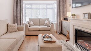 Un salon avec un canapé beige, une table basse sur laquelle sont posés des magazines, une cheminée et un téléviseur fixé au mur.