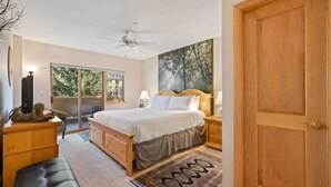 Une chambre à coucher comprenant un lit, des tables de chevet, un ventilateur de plafond, une grande fenêtre et une porte.