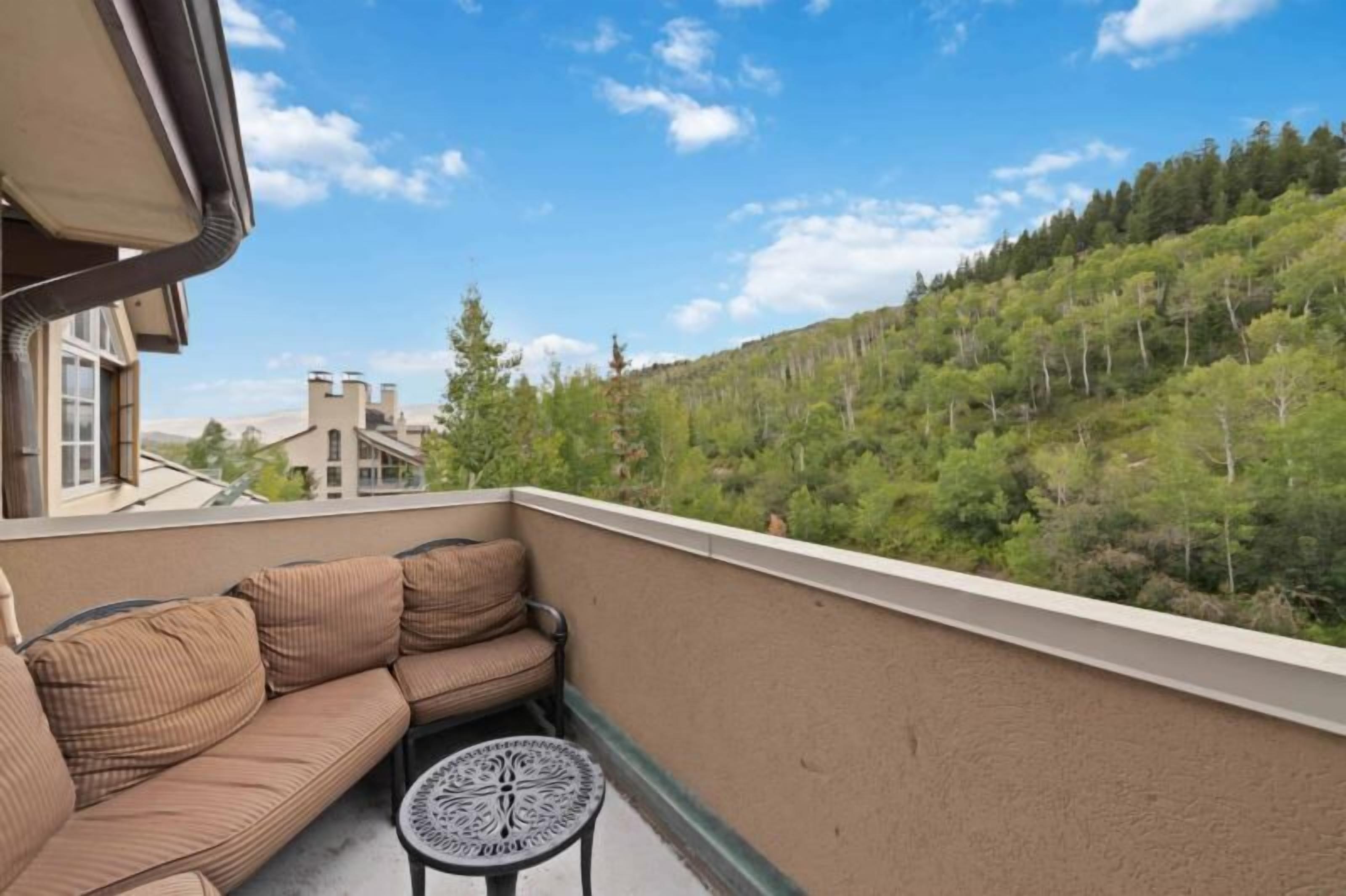 A balcony with a beige sofa and a small round table, overlooking a forested hillside.