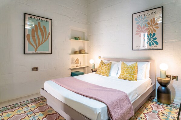 Une chambre d’hôtel comprenant un lit, une table de chevet, une étagère avec des objets décoratifs et deux œuvres d’art accrochées au mur.