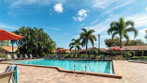 A swimming pool with lounge chairs and umbrellas, surrounded by palm trees and a clear sky. - Renovated Beachwalk 3 Bedroom Condo -5 minute walk to Beach! (Naples)