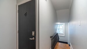 Un pasillo con una puerta oscura, una escalera de madera y un ventanal al final.