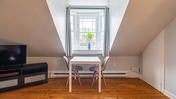 Una habitación con televisión, una mesa blanca con dos sillas, una ventana con cortinas y una planta.