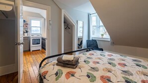 Un dormitorio con cama, una silla, un ventanal y vista a una cocina.