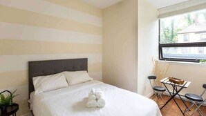 A hotel room with a bed, a small table with chairs, a window with a view, and a plant on a side table. - Quaint Loft with AC, Abundance of Sunlight (Halifax)