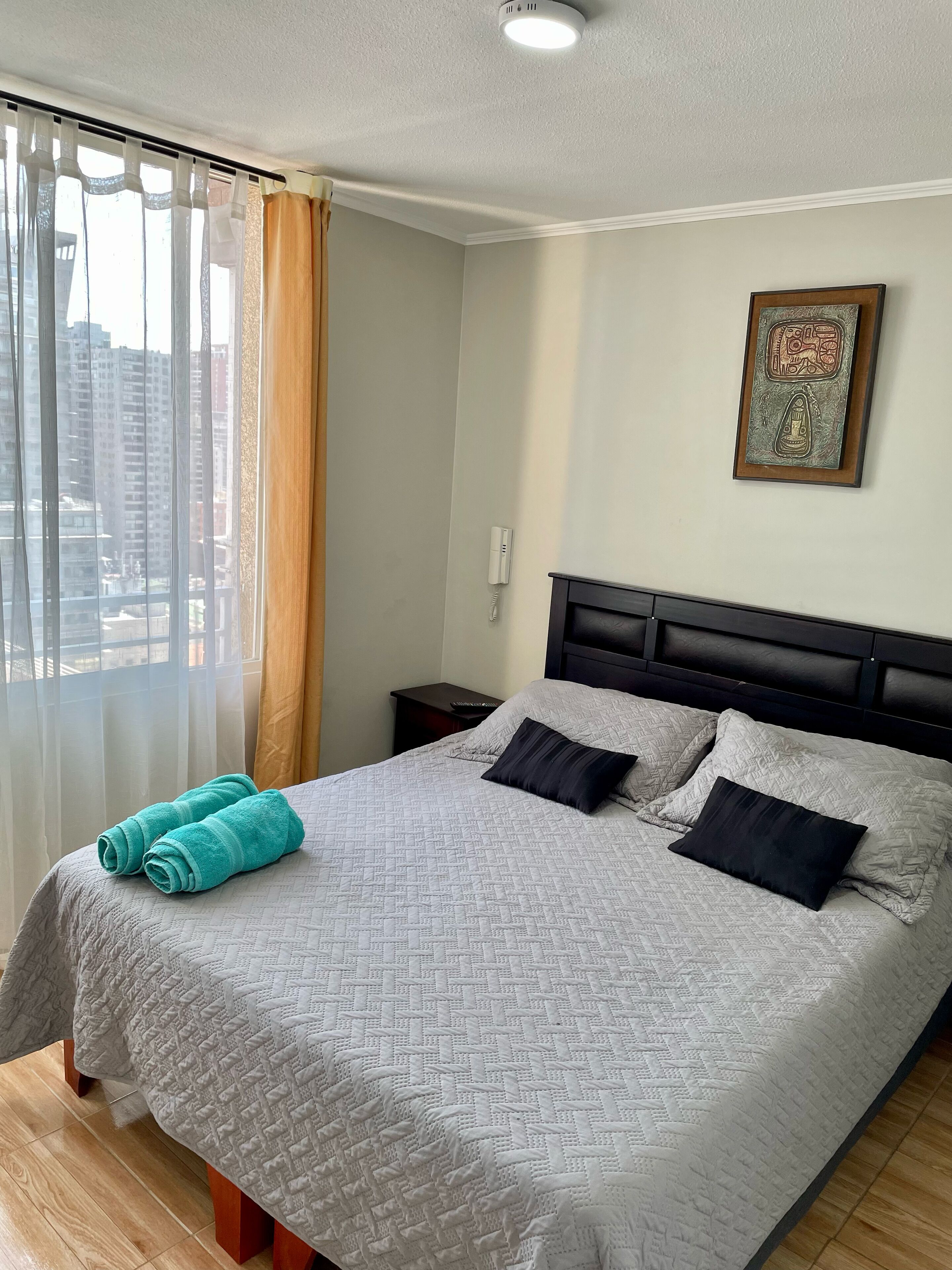 A neatly made bed with a patterned bedspread and two pillows, a bedside table with a phone, and a window with sheer curtains.