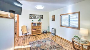A room with wooden flooring, a flat-screen TV mounted on the wall, a bookshelf, a chair, a window with blinds, and a table with a lamp.