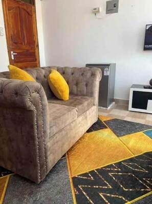 A grey tufted sofa with yellow cushions, a black and yellow patterned rug, and a white wall with a thermostat.
