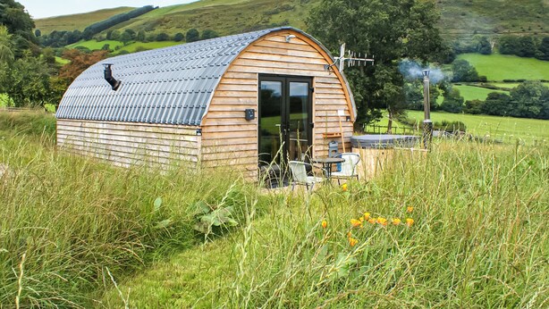 A wooden barn with a curved roof, a door, and a chimney emitting smoke, surrounded by a grassy field and hills. - Creyr Glas Pod -UK41796 (Penybontfawr, near Llanfyllin)