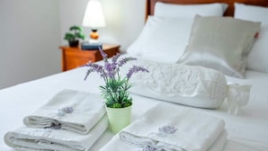 A neatly made bed with white linens and lavender towels, a small potted plant with purple flowers, and a bedside table with a lamp and a book.