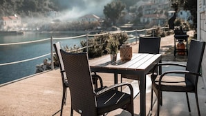 An outdoor patio with wicker chairs and a table, overlooking a body of water and a mountainous landscape.