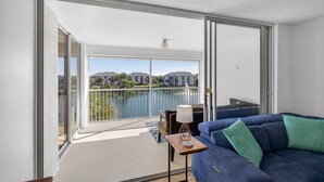 A modern living room with a blue sofa, a small table with a lamp, and a view of a lake and buildings through large glass doors. - Beach Access Resort Condo with Breath-Taking View (Jupiter)