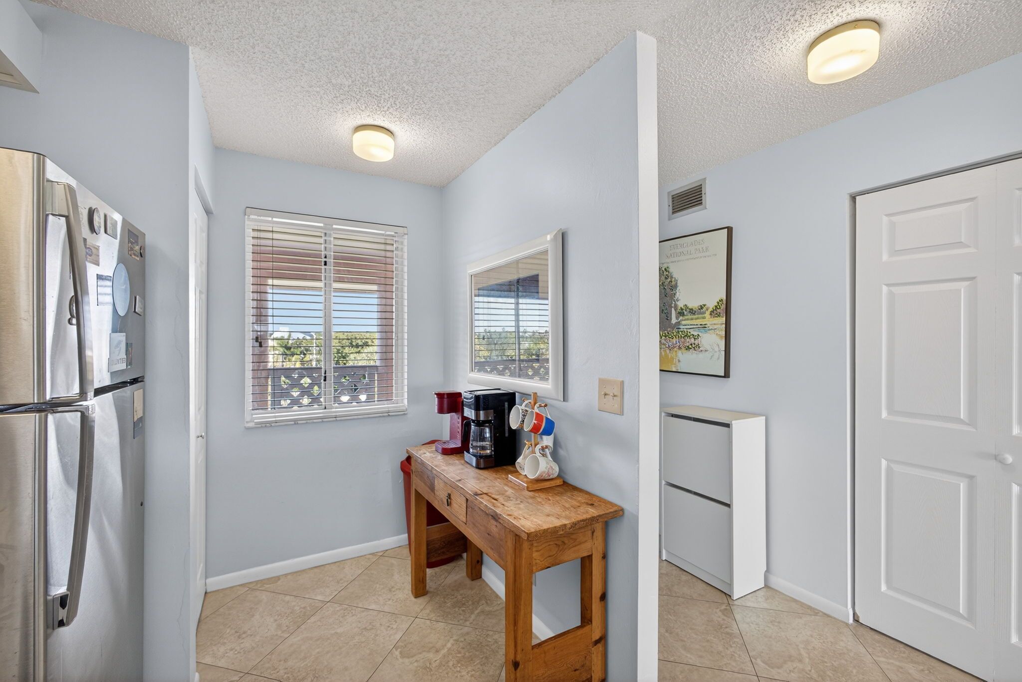 A compact kitchen with a refrigerator, a wooden table with a coffee maker, and a window with blinds.