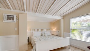 Ein Schlafzimmer mit einem weißen Bett, einem Nachttisch, einer Wand-Klimaanlage und einem Fenster mit Jalousien.