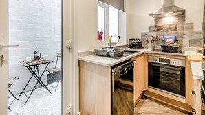 Une cuisine compacte équipée d’un lave-vaisselle, d’un four et d’un grille-pain. Une fenêtre offre une vue sur l’extérieur, et une petite table est agrémentée d’une machine à café et de tasses.