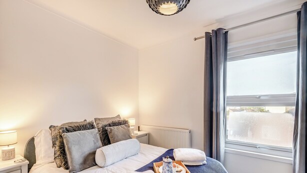 Un lit bien fait, recouvert d’une couverture à rayures bleues et blanches, un plateau en bois avec une théière, et un tapis blanc et moelleux.