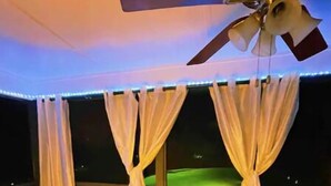 Una stanza con ventilatore a soffitto, tende bianche, un letto, un comodino e vista sul cielo notturno.