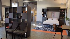 A modern hotel room with a bed, a desk, a bookshelf, and a seating area. - Suite with AC, Industrial Styling (Bedford)