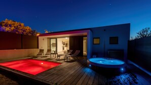 Een moderne buitenzwembadzone met een jacuzzi, een houten terras en ligbedden.