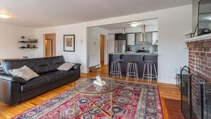 A modern living room with a black leather sofa, a glass coffee table, and a patterned rug. There is a kitchen area with a brick fireplace and a bar stool. - Beautiful & Spacious Apt, AC (Bedford)
