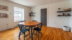 A room with a wooden floor, a round table with chairs, a window with blinds, and shelves with decorative items. - Beautiful & Spacious Apt, AC (Bedford)